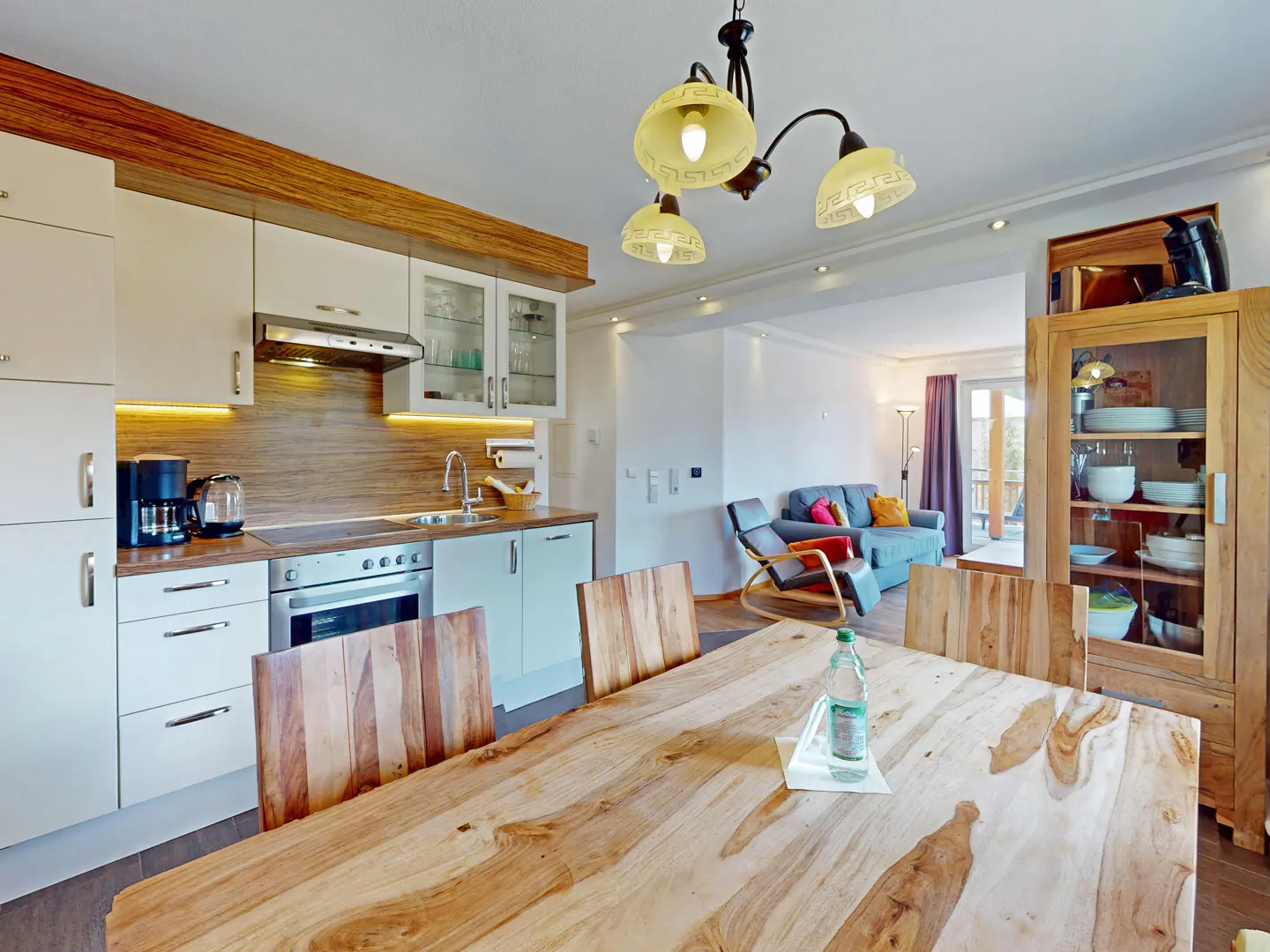 A bright, modern kitchen and dining area with a large wooden table and chairs. White cabinets and stainless steel appliances are visible. A blue sofa is in the background.