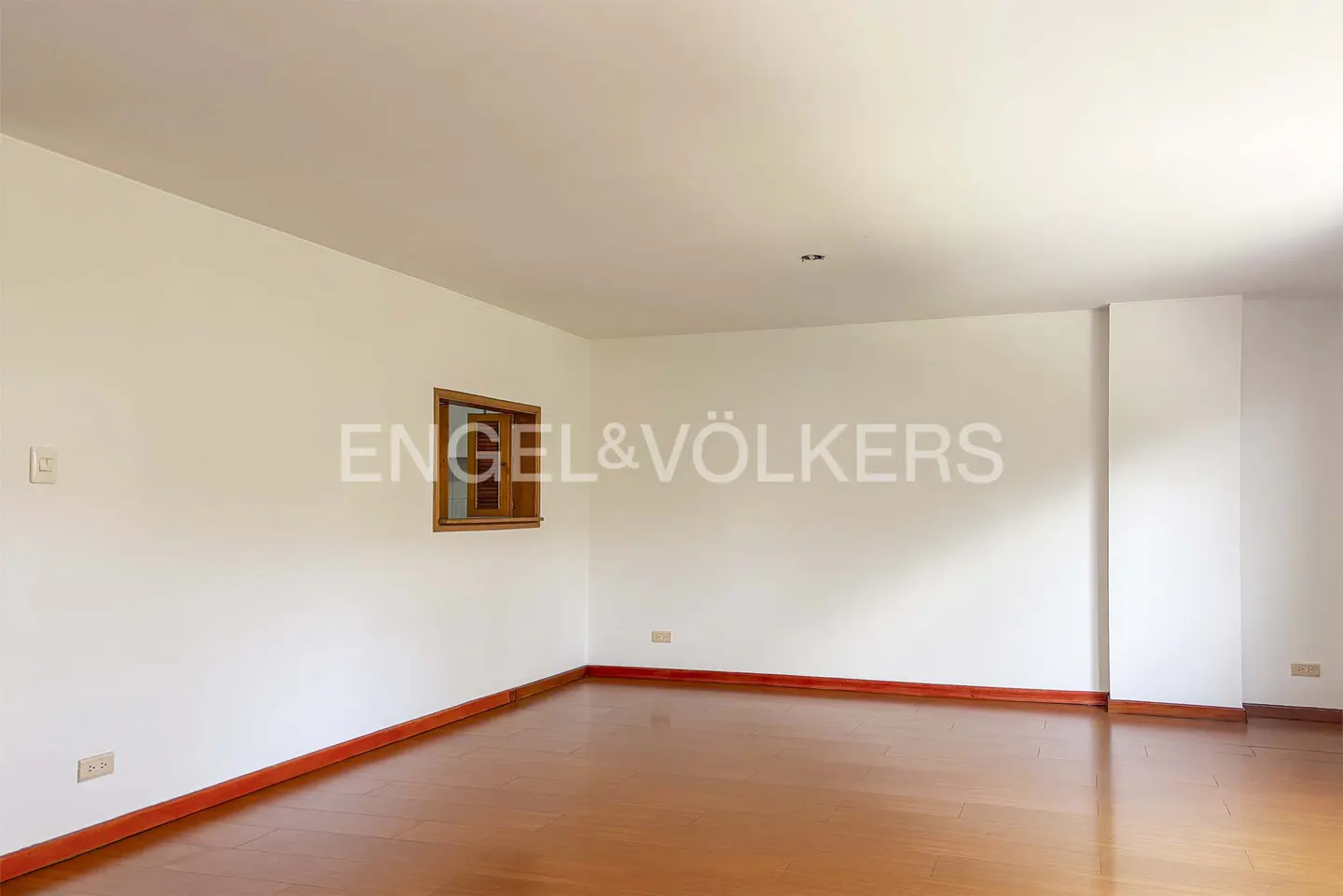 Empty room with hardwood floors, white walls, and a small window. Engel & Völkers logo on the wall.