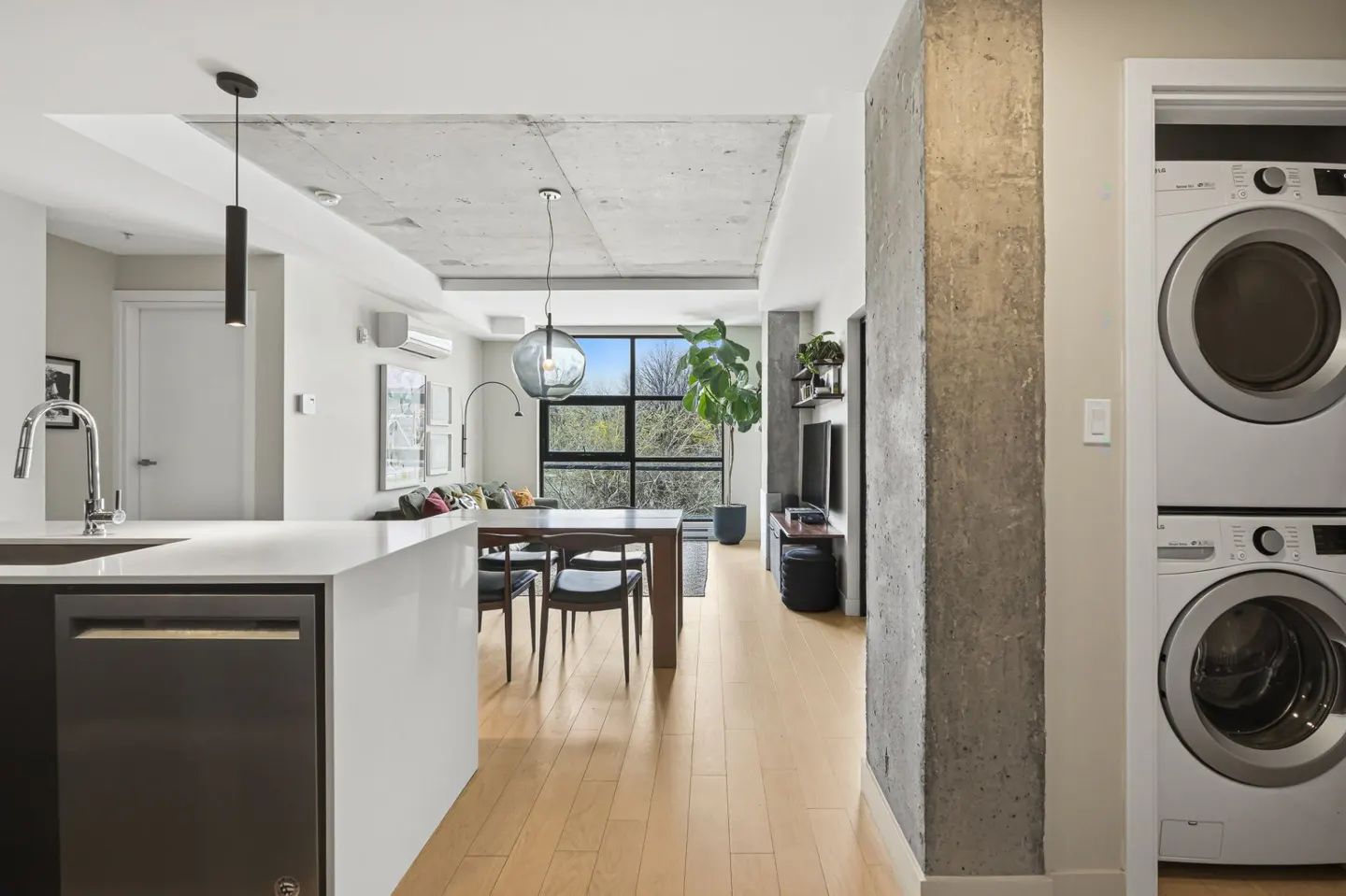 A modern apartment interior with a kitchen island, dining table, and living area. Concrete ceilings and a pillar add an industrial touch. Washer and dryer are visible.