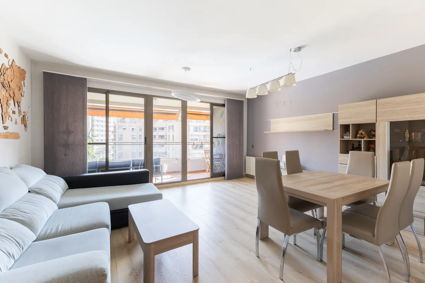Bright living room with a gray sofa, wood table, and chairs. Sliding glass doors lead to a balcony with city views. A wooden world map hangs on the wall.