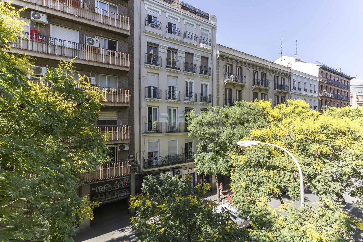 View of a street in a city with multi-story apartment buildings, balconies, and lush green trees under a clear blue sky.
