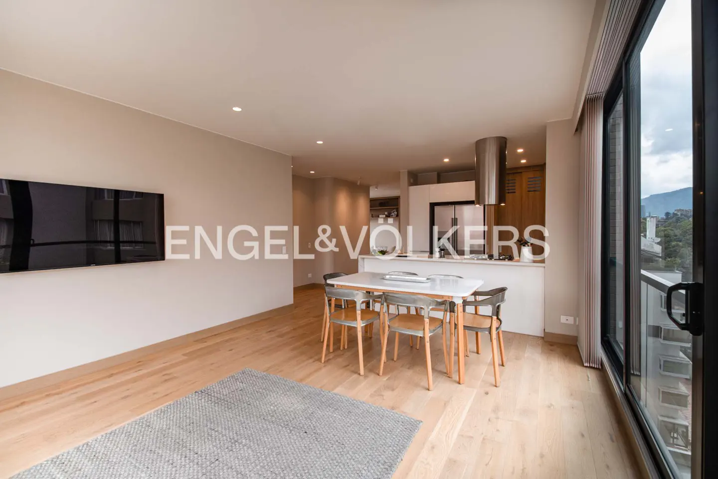 A modern apartment interior with a white dining table, wooden chairs, and a gray rug on a light wood floor. A kitchen and large windows are visible.