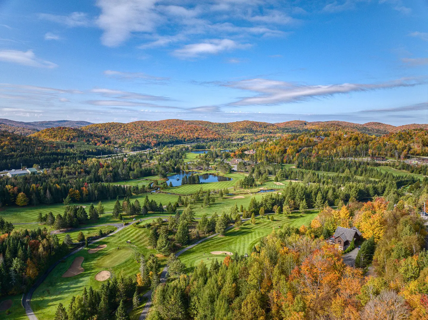Aerial view of a lush green golf course with ponds, surrounded by colorful autumn trees and rolling hills under a blue sky.