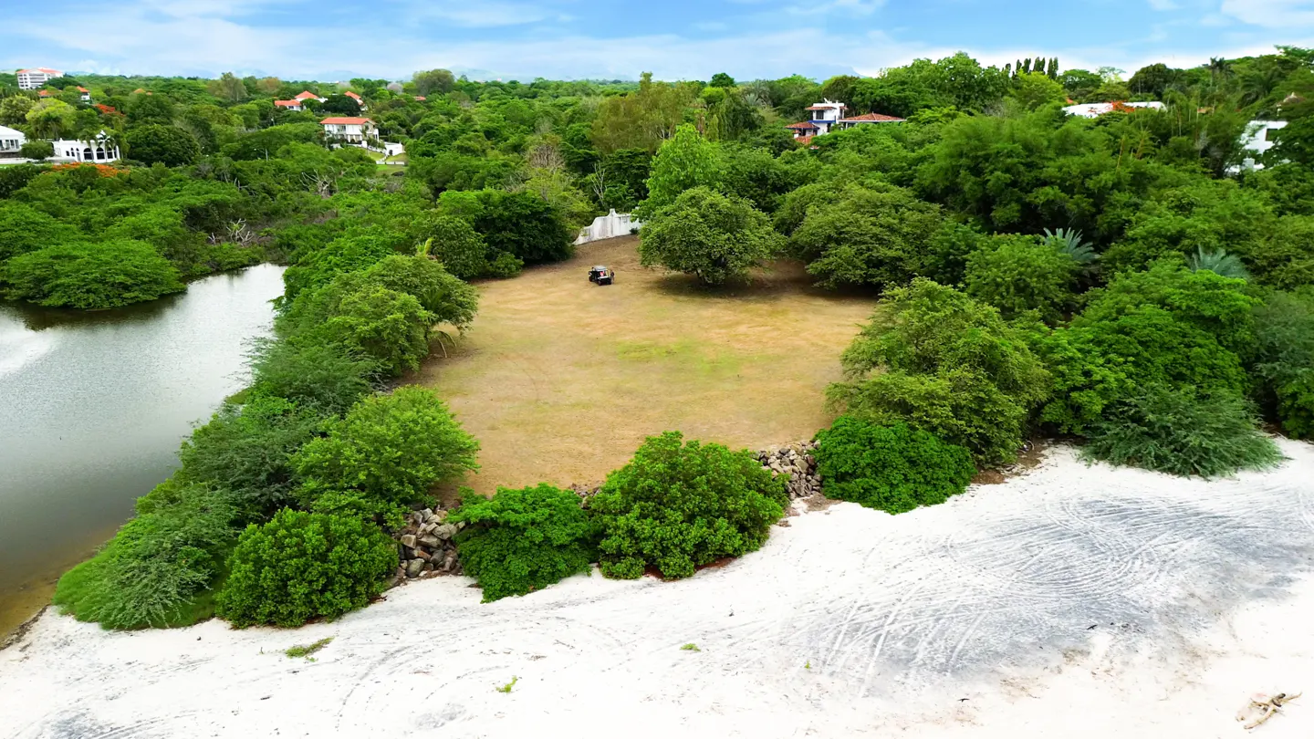 Aerial view of a vacant lot with green trees, a lake, and a sandy beach under a blue sky.