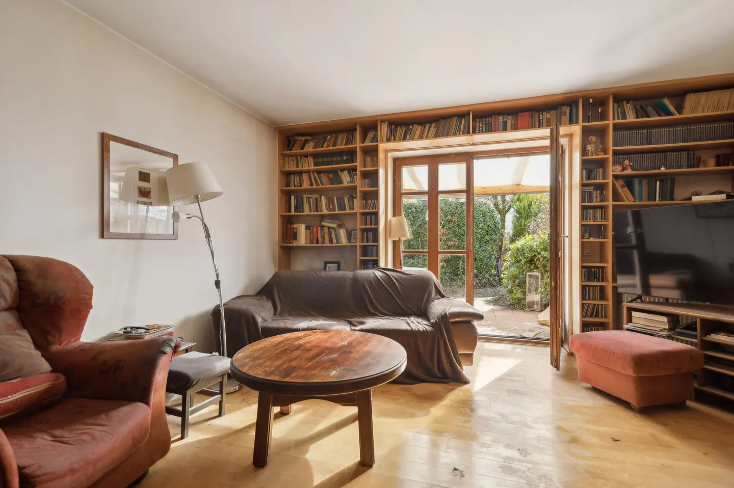 Living room with hardwood floors, a brown sofa, a red armchair, and floor-to-ceiling bookshelves. A glass door leads to a green garden.