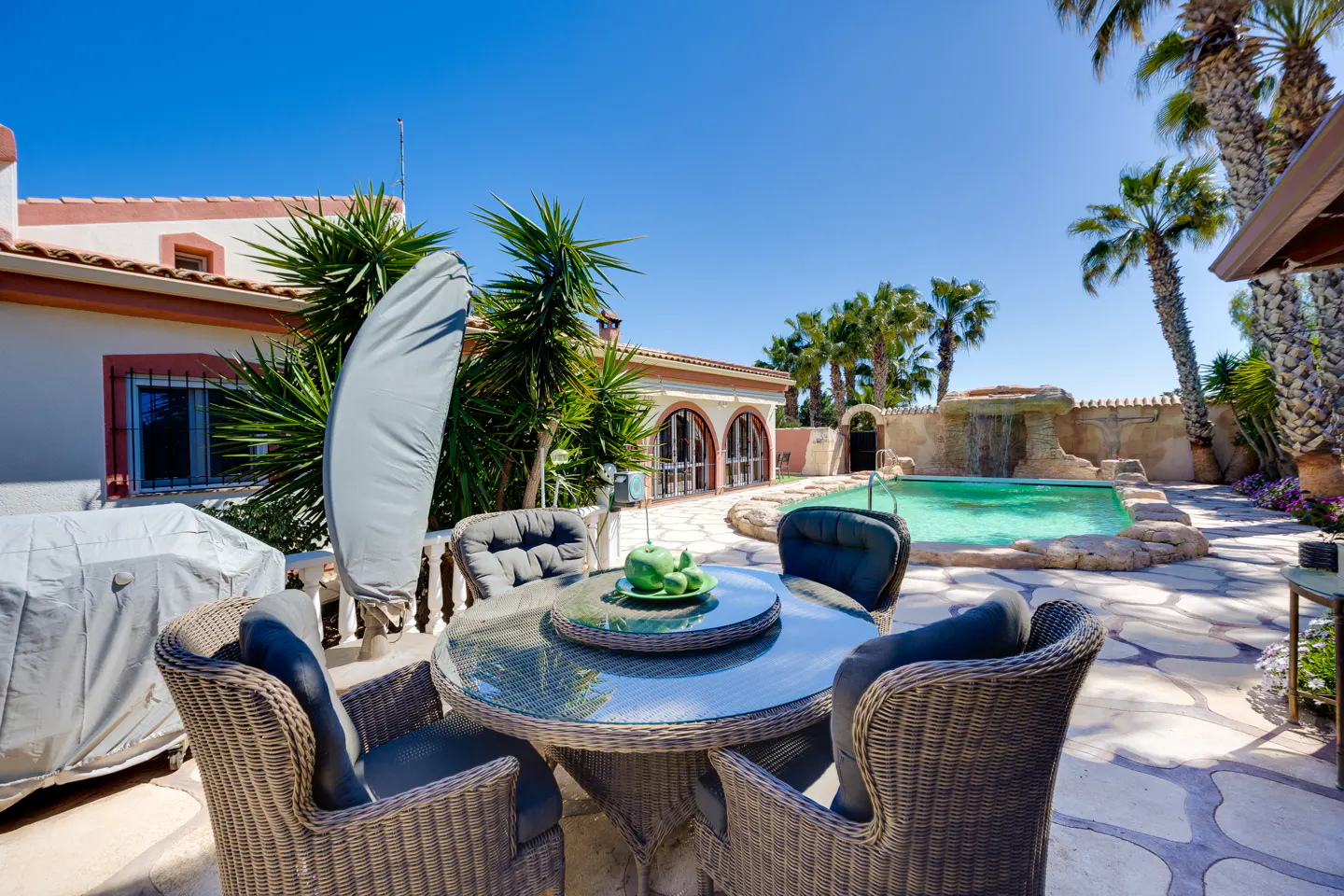 Outdoor patio with wicker furniture, a glass table, and a pool with a waterfall feature in the background on a sunny day.
