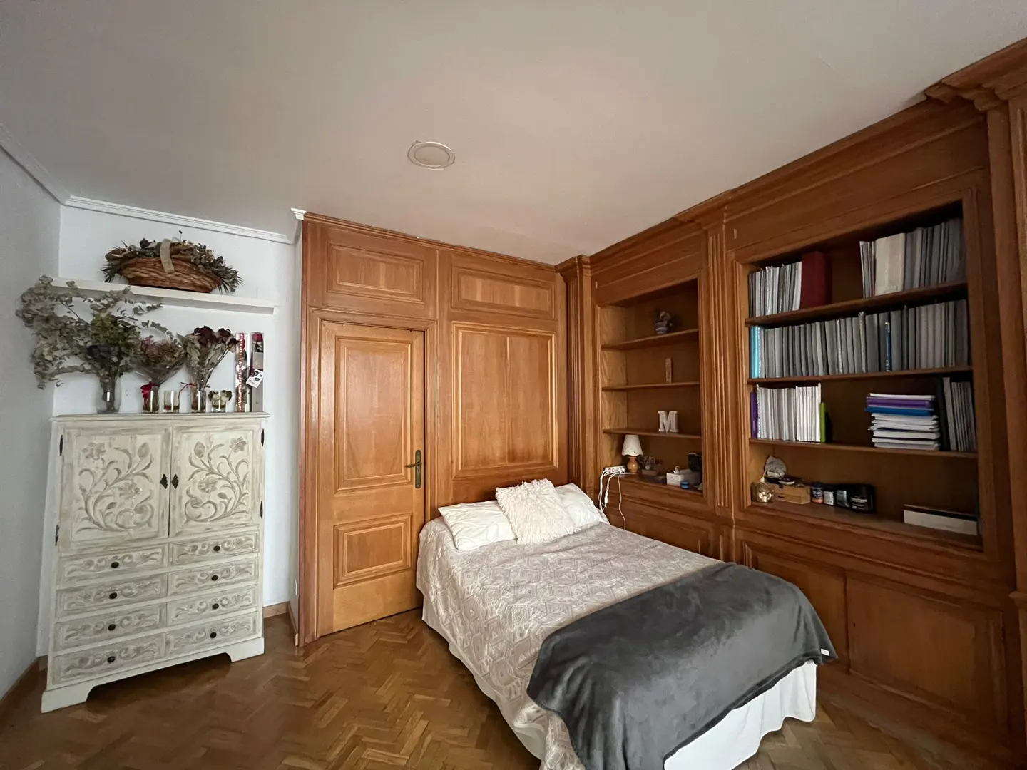 Bedroom with a bed, built-in wooden bookshelves, a white carved dresser, and herringbone wood floors.