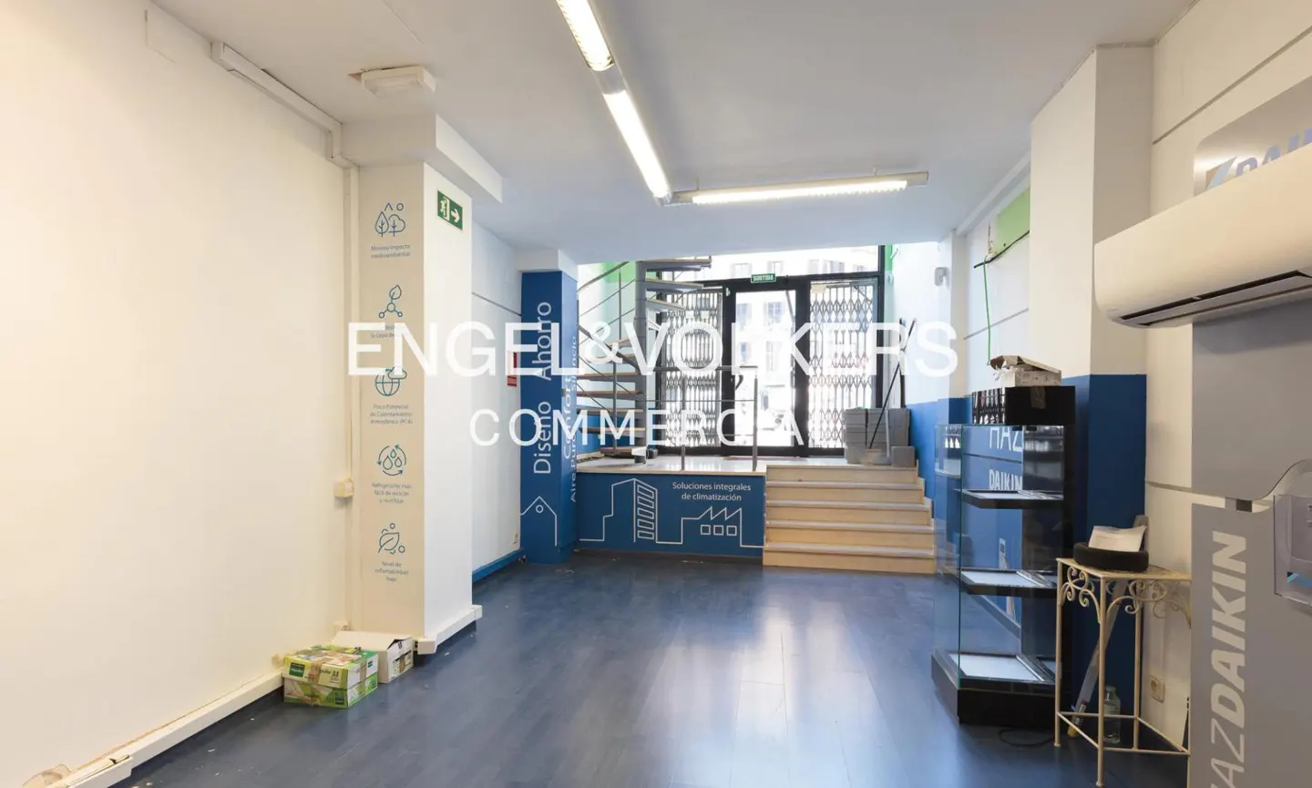Bright commercial space with blue floor, white walls, and Engel & Völkers branding. Stairs lead to a metal gate and glass door.