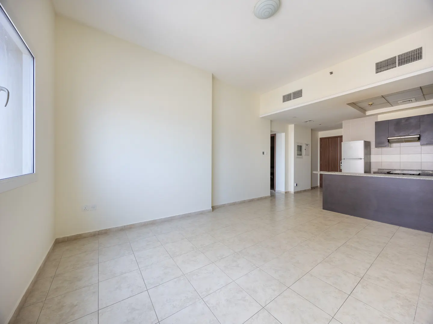 An empty apartment with beige walls and tile floors. A kitchen with gray cabinets and a breakfast bar is visible in the background.