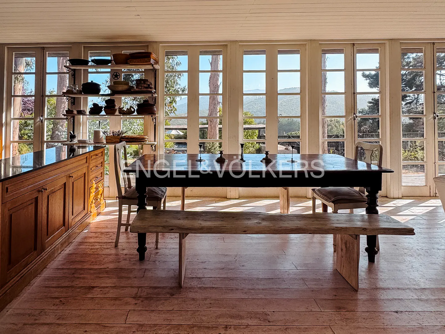 Bright dining room with a dark wood table, bench, chairs, and a view of trees and mountains through white-framed windows.
