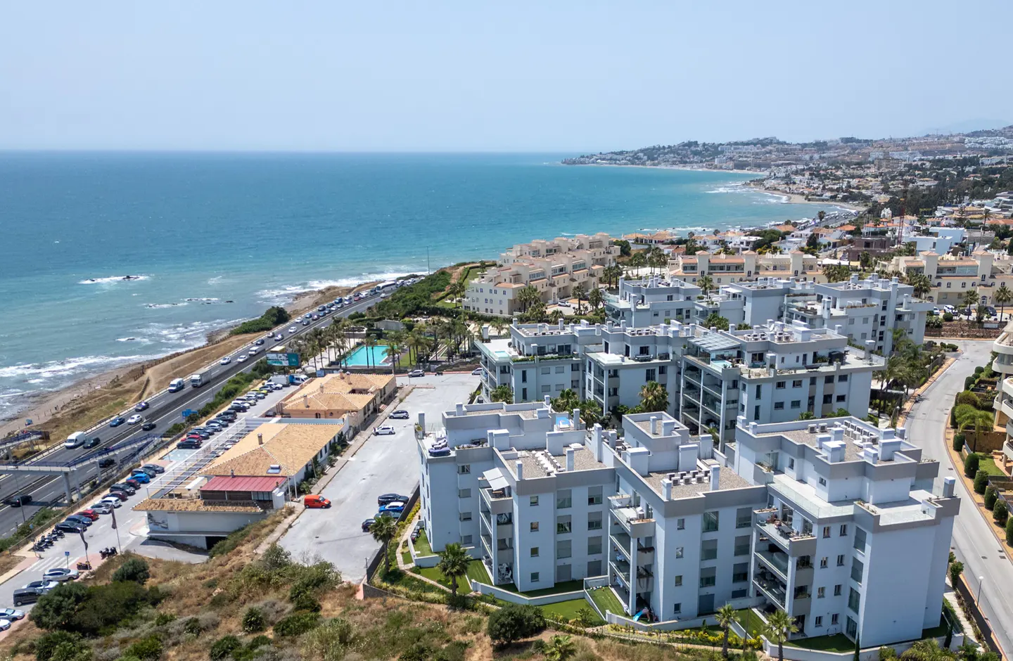 Aerial view of white apartment buildings near a blue ocean and beach, with a road and cars visible.