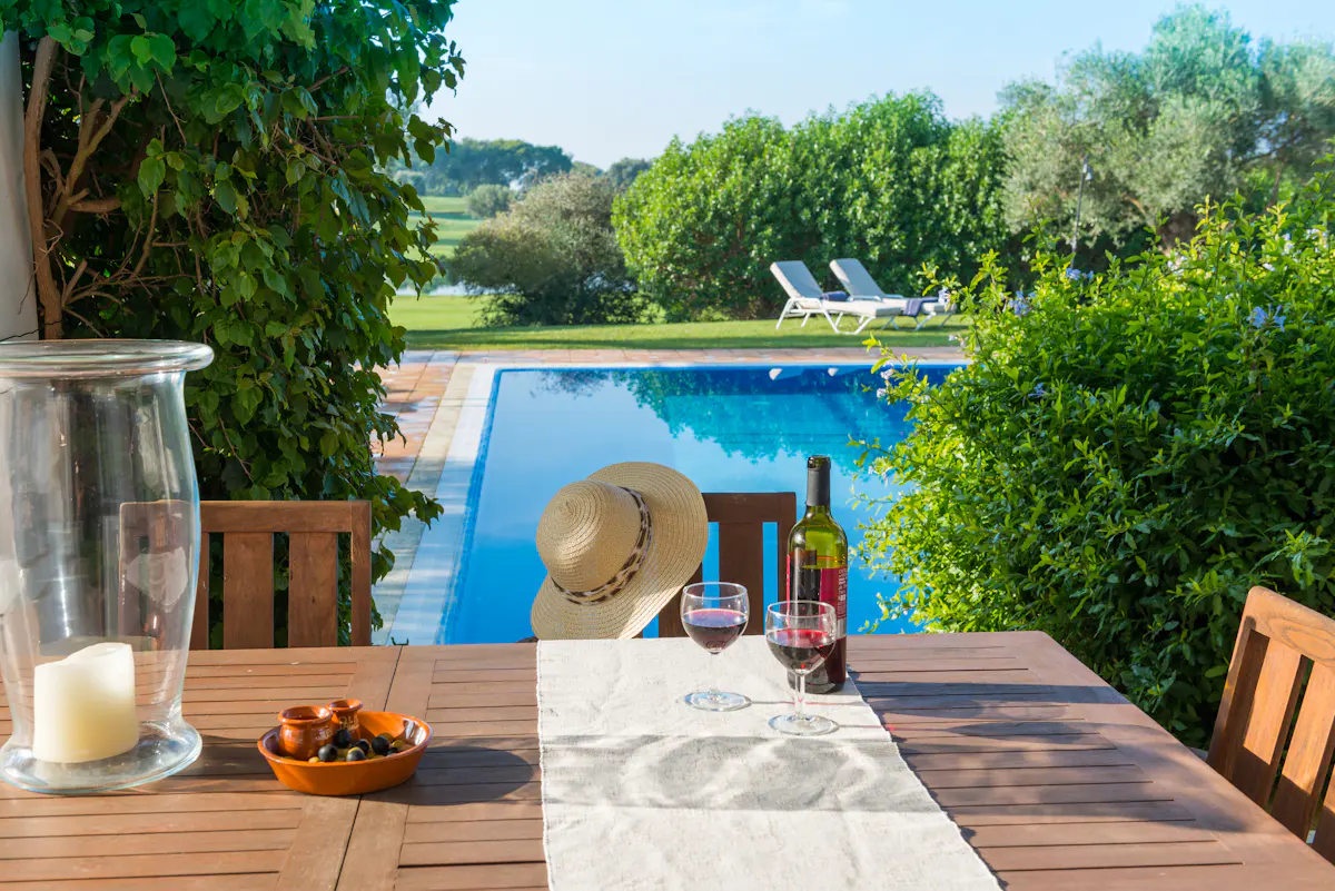 Outdoor dining table with wine, glasses, olives, and a hat, overlooking a blue pool and green landscape.