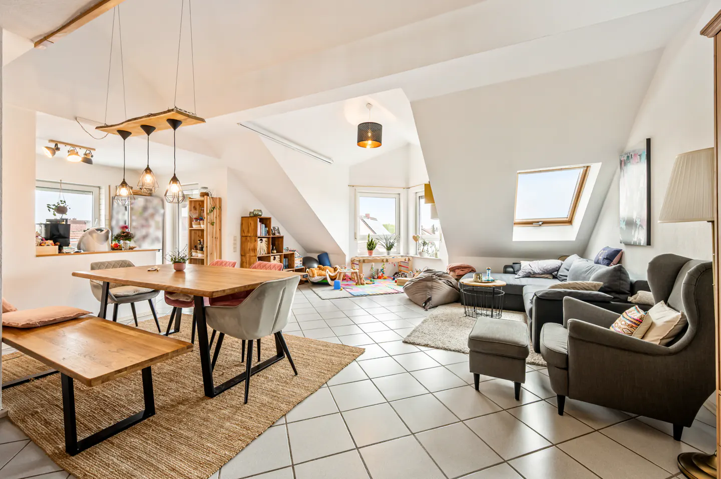 Bright, open-plan attic living area with a wooden dining table, grey armchairs, a sofa, and a children's play corner.