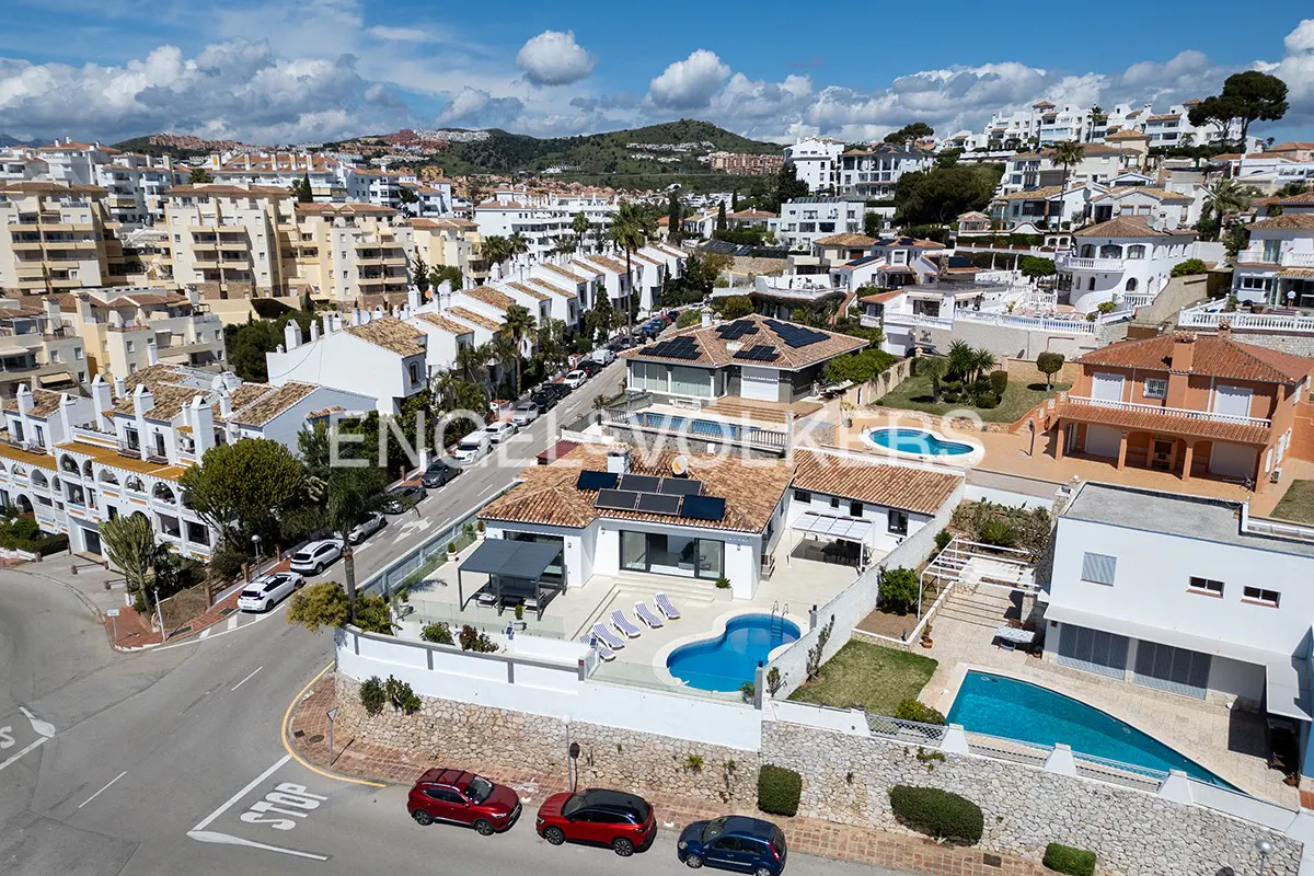 Aerial view of a Spanish neighborhood with white buildings, red tile roofs, and blue pools on a sunny day.