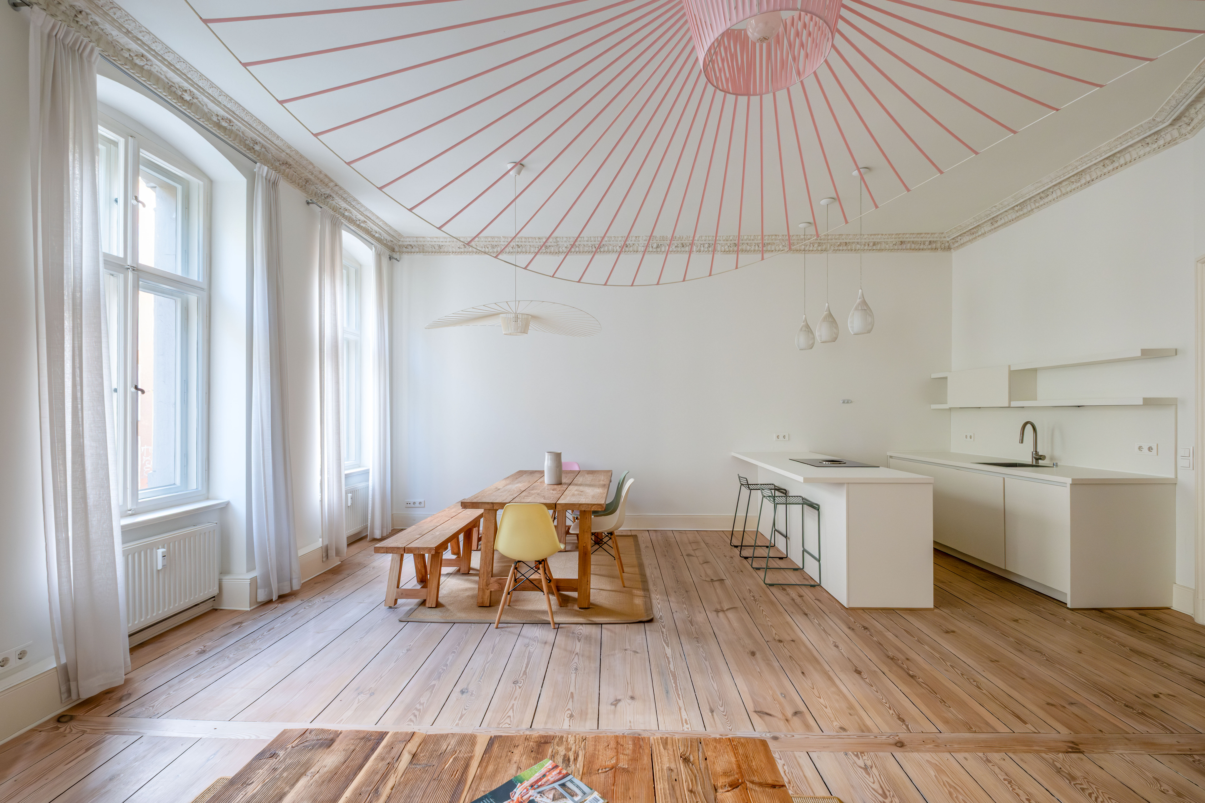 Modern dining area with wooden table, bench, yellow chair, white kitchen island, and a unique pink radial ceiling light fixture. [AI-generated text]