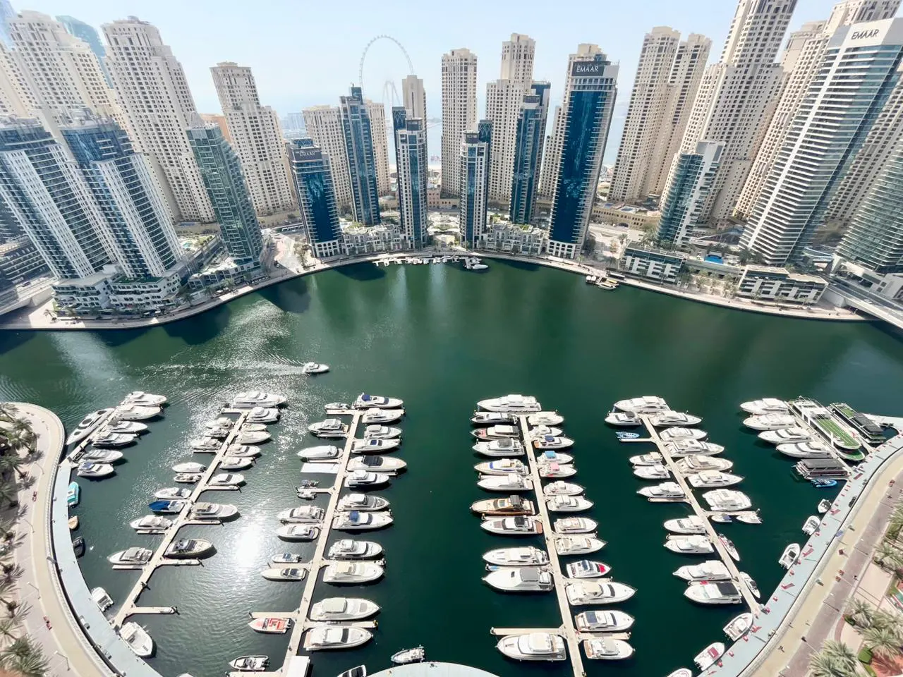 Aerial view of Dubai Marina with yachts docked in the harbor, surrounded by modern high-rise buildings under a clear sky.