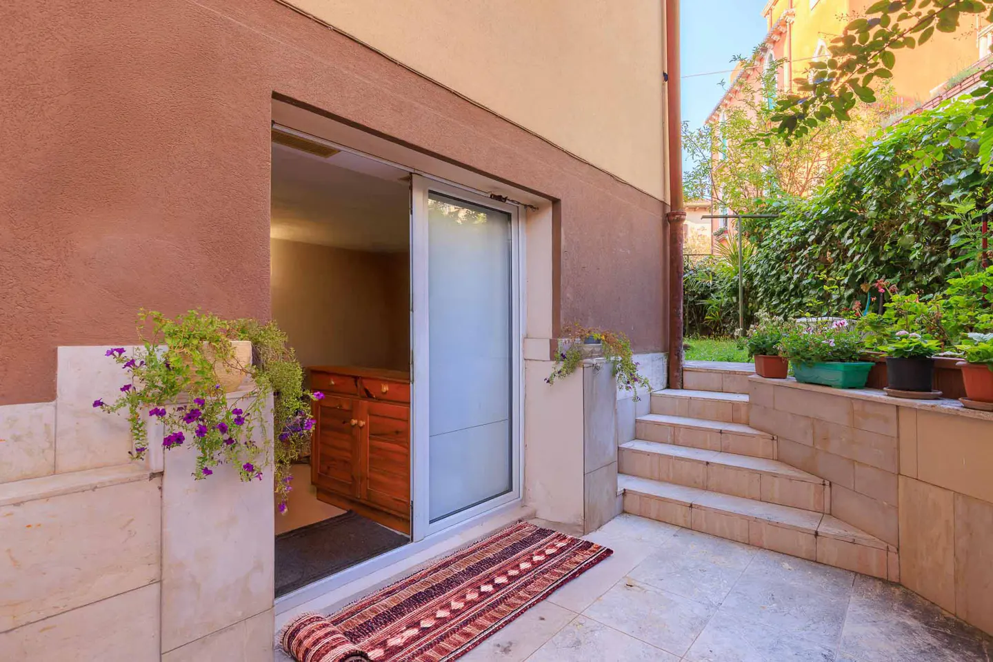 Exterior view of a home with a sliding glass door, a rolled rug, and steps leading to a garden. The walls are brown and beige.