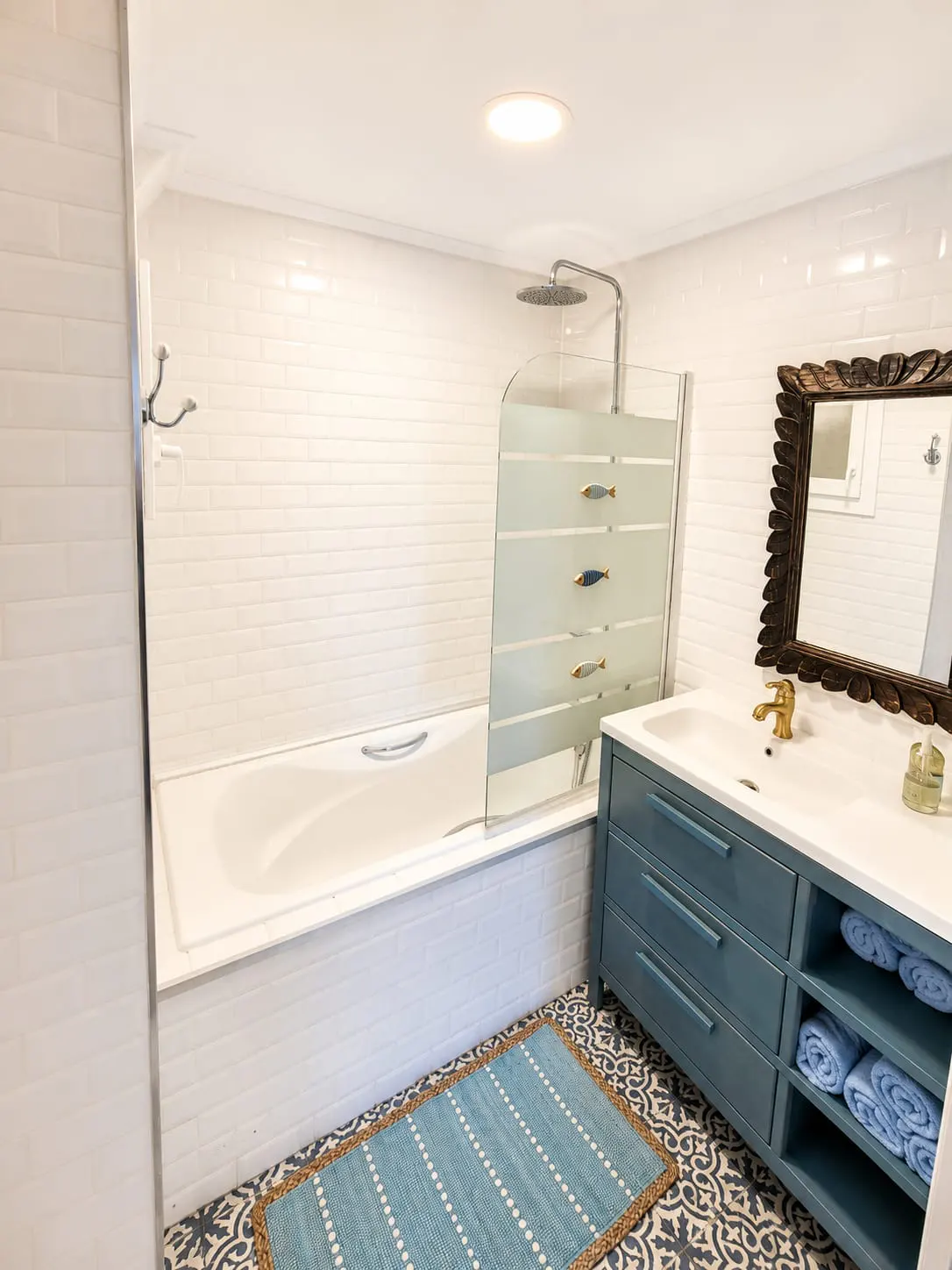 Bathroom with white subway tile, blue vanity, and tub with a glass screen decorated with fish. A blue rug sits on a patterned tile floor.