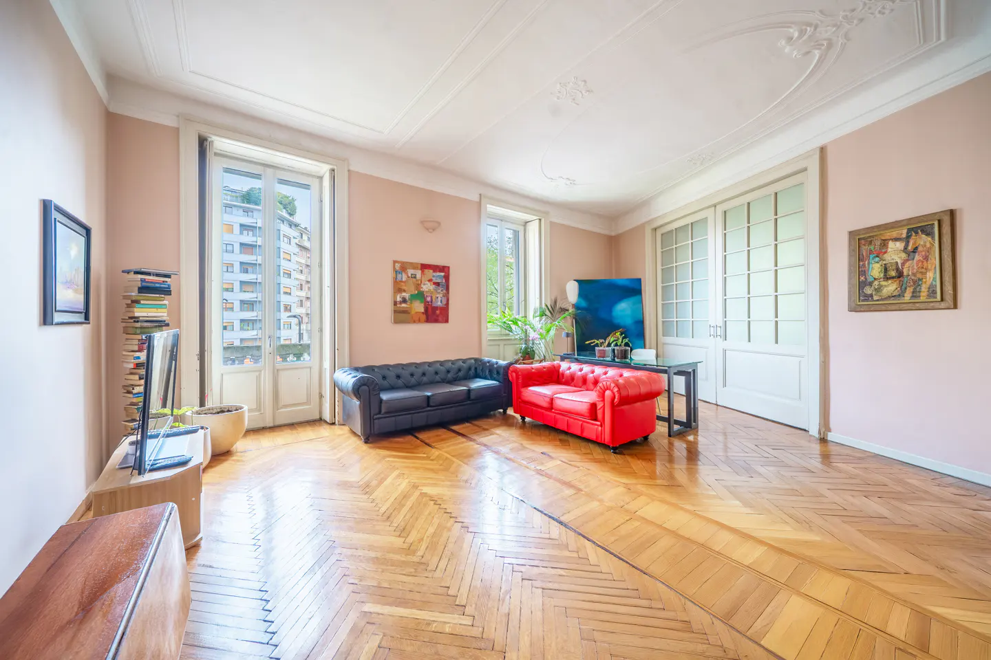A bright living room with herringbone wood floors, a black leather sofa, and a red leather sofa. There are paintings on the walls.