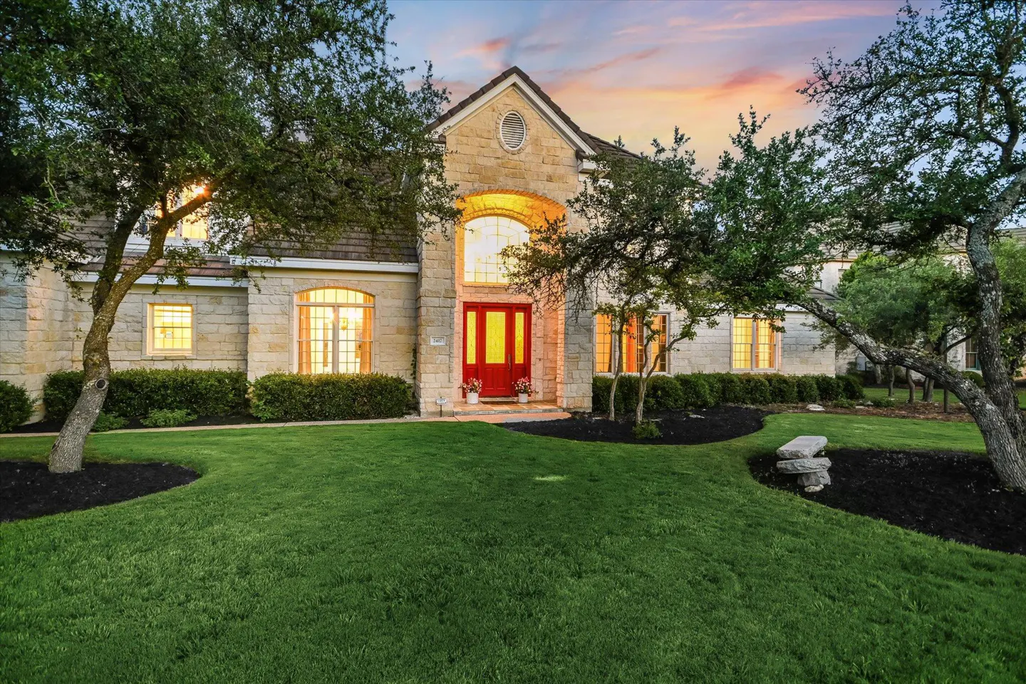 Stone house with red door, green lawn, and trees. Windows glow with warm light. Sunset sky in the background.
