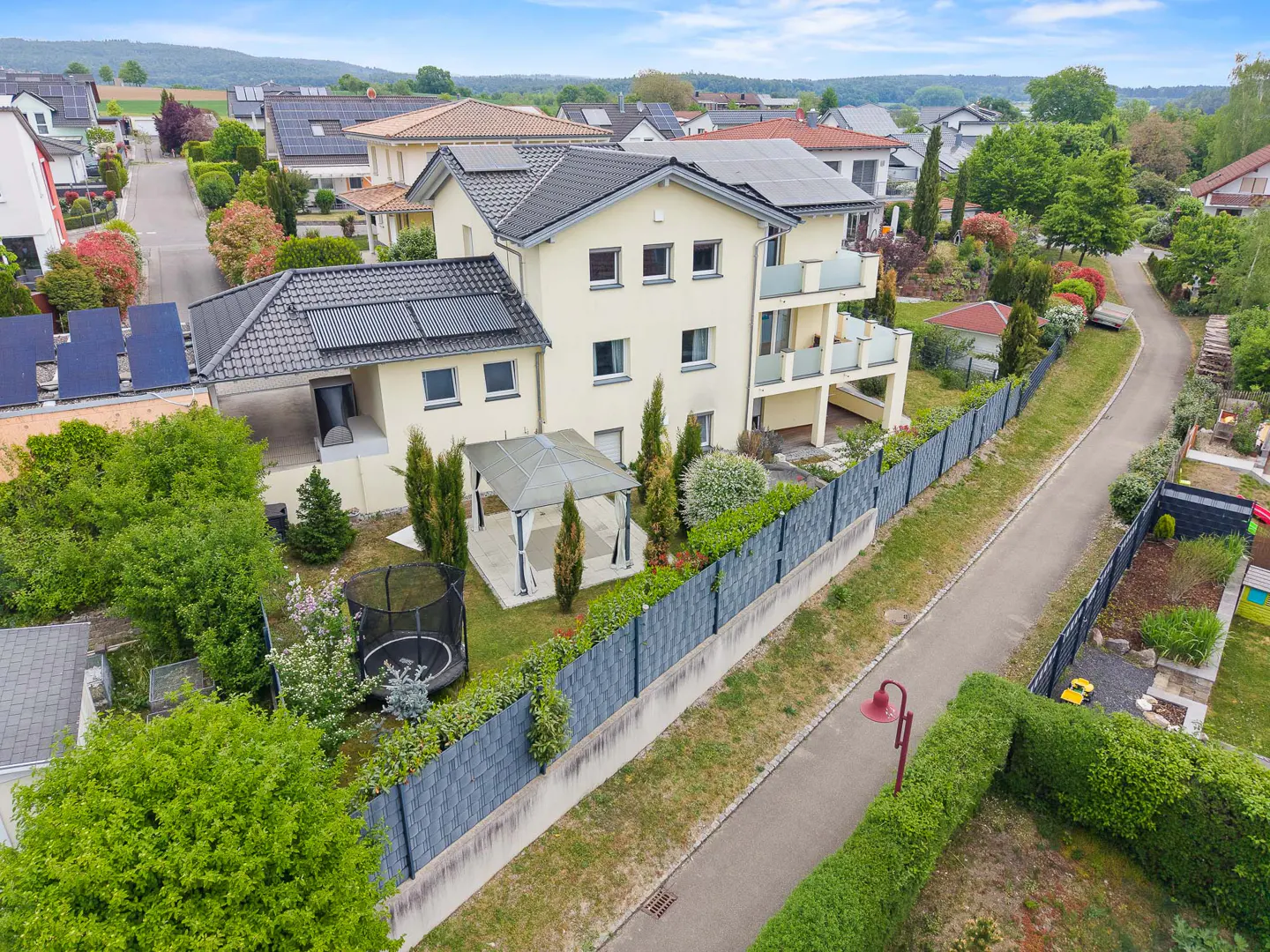 Aerial view of a two-story beige house with a gray roof, solar panels, a gazebo, and a trampoline in the backyard.