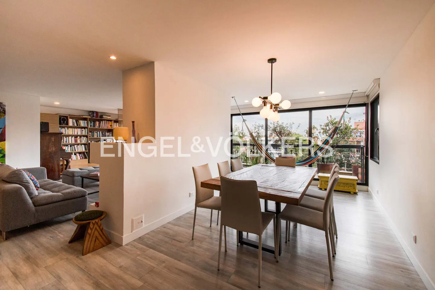 Open-concept living space with a dining table, chairs, and a hammock by the window. A gray sofa and bookshelf are visible in the background.