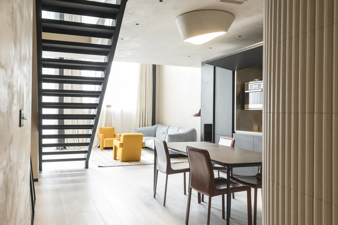 A modern apartment interior with a black staircase, dining table, yellow chairs, and a gray sofa.