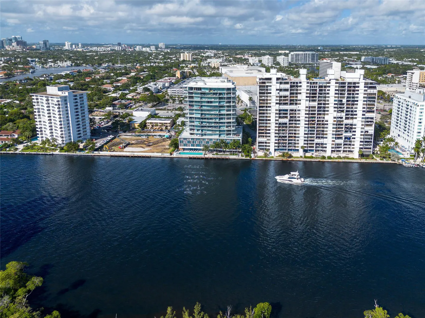 Aerial view of white condos along a dark blue waterway, with a white boat moving across the water. City skyline in the background under a partly cloudy sky.