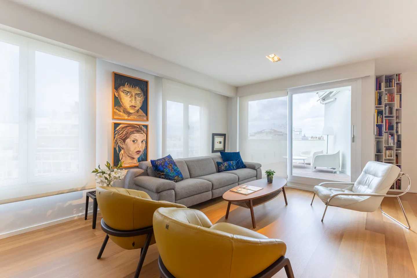 Bright living room with wood floors, gray sofa with blue pillows, yellow chairs, and art on the wall. Sliding glass doors lead to a balcony.