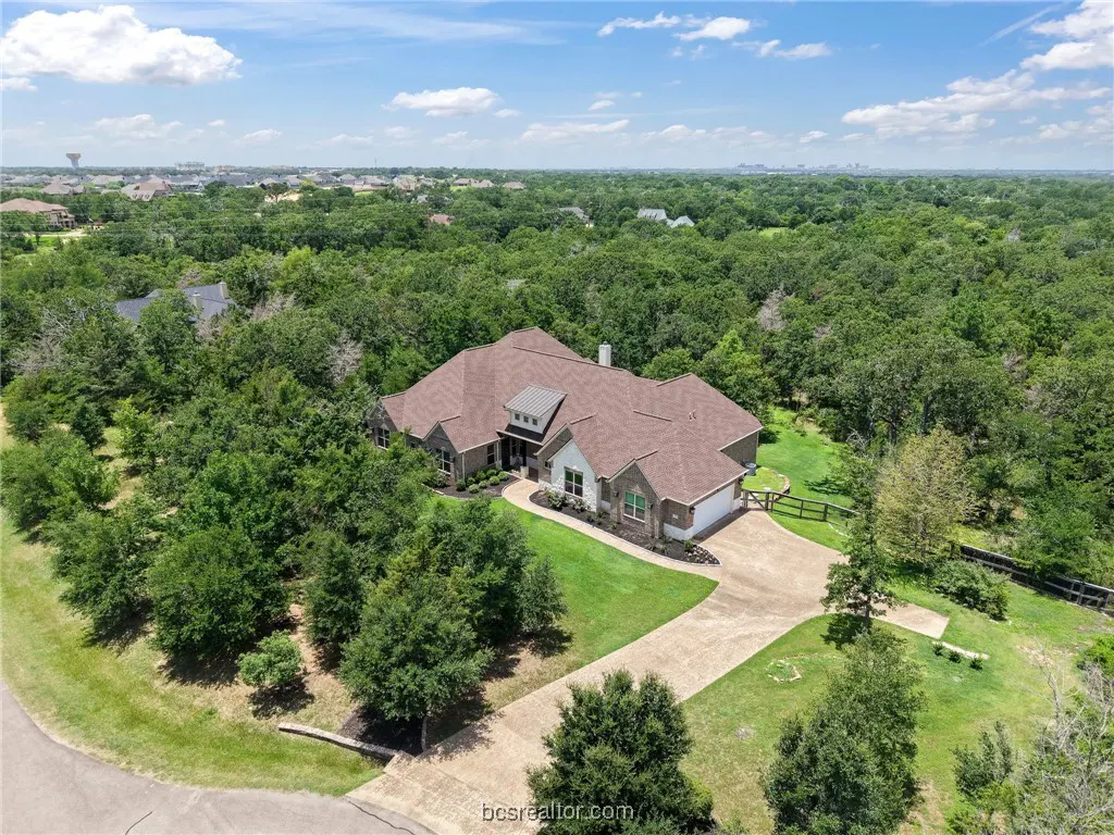 Aerial view of a large, single-story brick house with a brown roof, surrounded by lush green trees and a long driveway. Blue sky with clouds above.