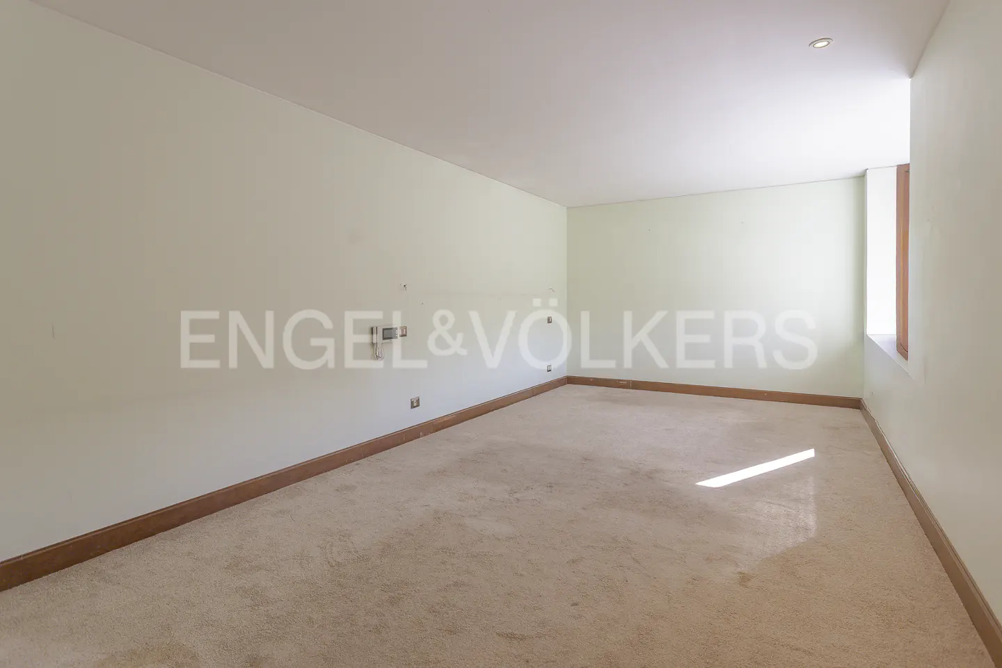 Empty room with beige carpet, light green walls, and white ceiling. Engel & Völkers logo on the wall. Window on the right.
