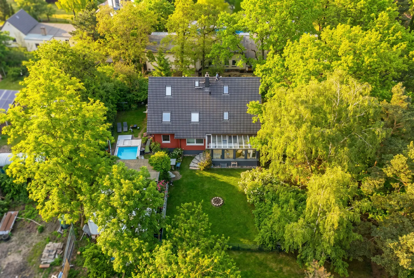 Aerial view of a red house with a dark roof, surrounded by green trees, a pool, and a fire pit in the backyard.