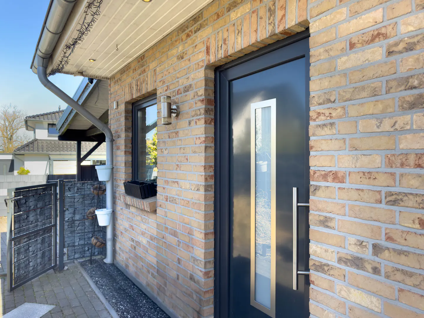 Exterior shot of a brick house with a dark gray front door with a vertical glass panel and a silver handle. A window with a black planter box is to the left of the door.