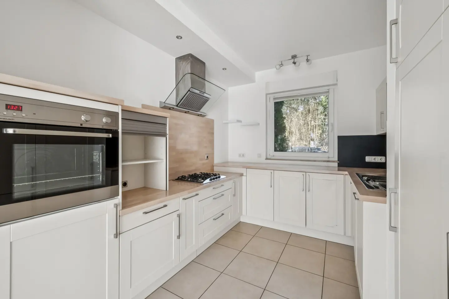 Bright kitchen with white cabinets, light wood countertops, stainless steel oven and range hood, and beige tile flooring. A window overlooks trees.