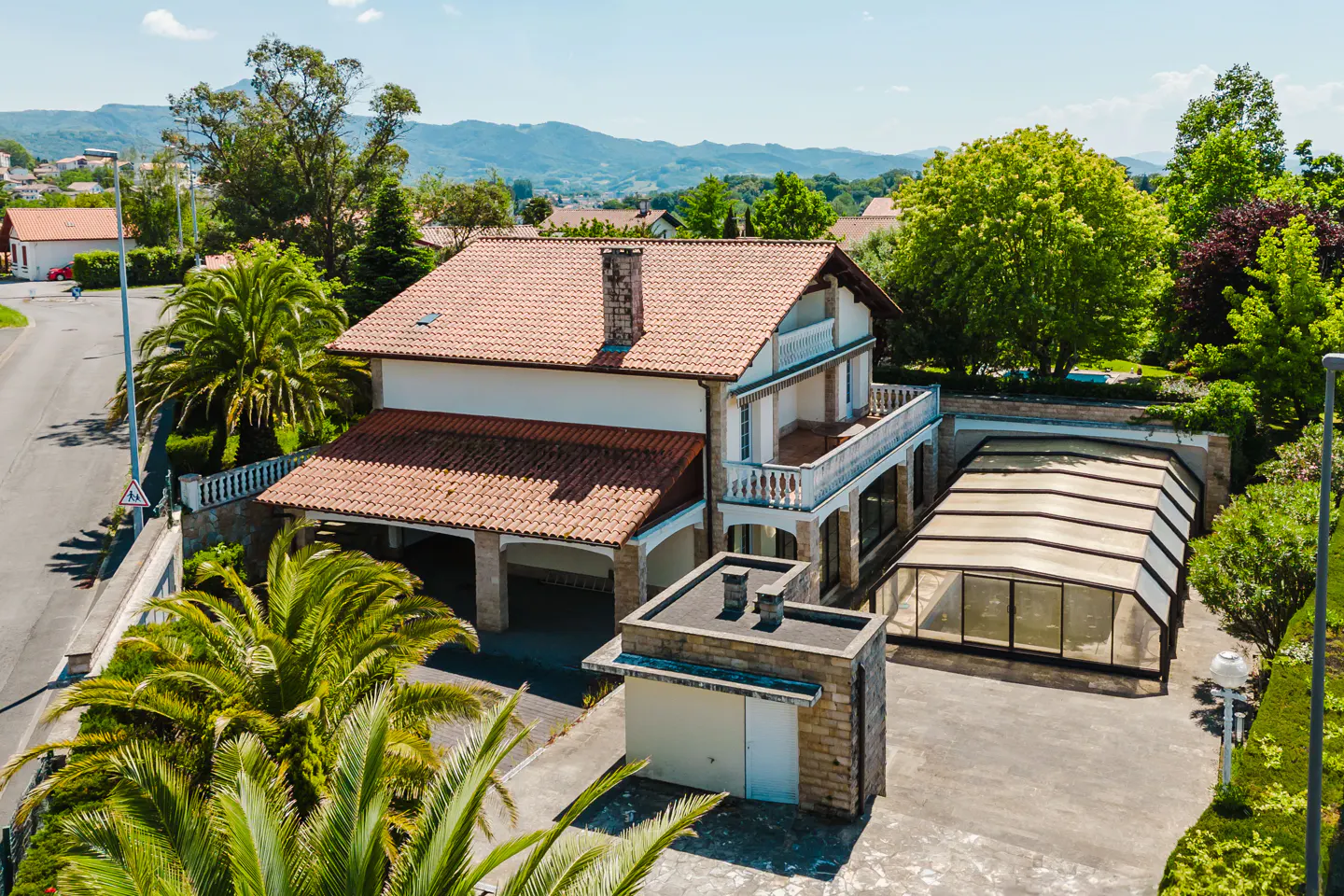Aerial view of a two-story white house with a red tile roof, a balcony, a covered pool, and palm trees.
