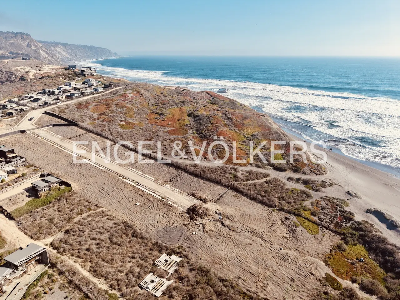 Aerial view of coastal real estate with ocean backdrop. Undeveloped land with sparse vegetation and sandy beach. Blue sky.
