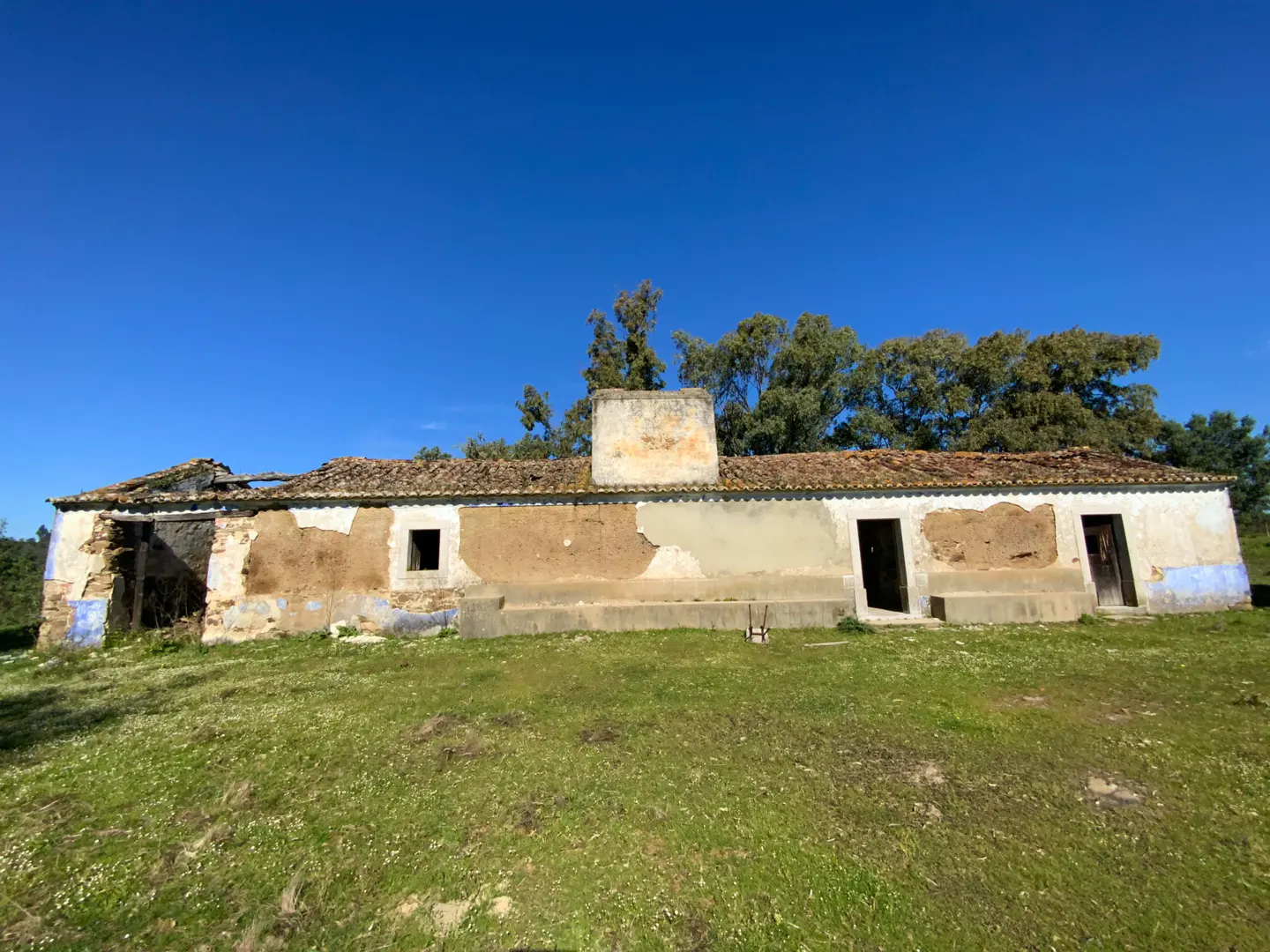 Exterior view of a dilapidated, one-story farmhouse with a brown tile roof, crumbling walls, and a chimney, set against a clear blue sky and green field.