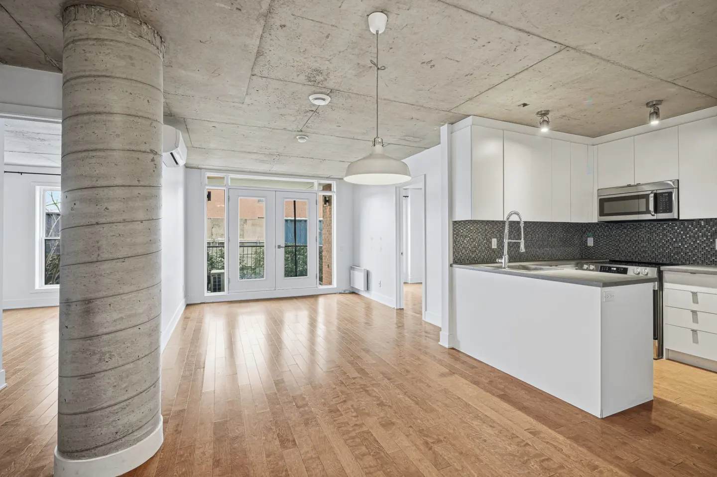 Bright, modern apartment with hardwood floors, concrete pillar, and white kitchen. Double doors lead to a balcony.
