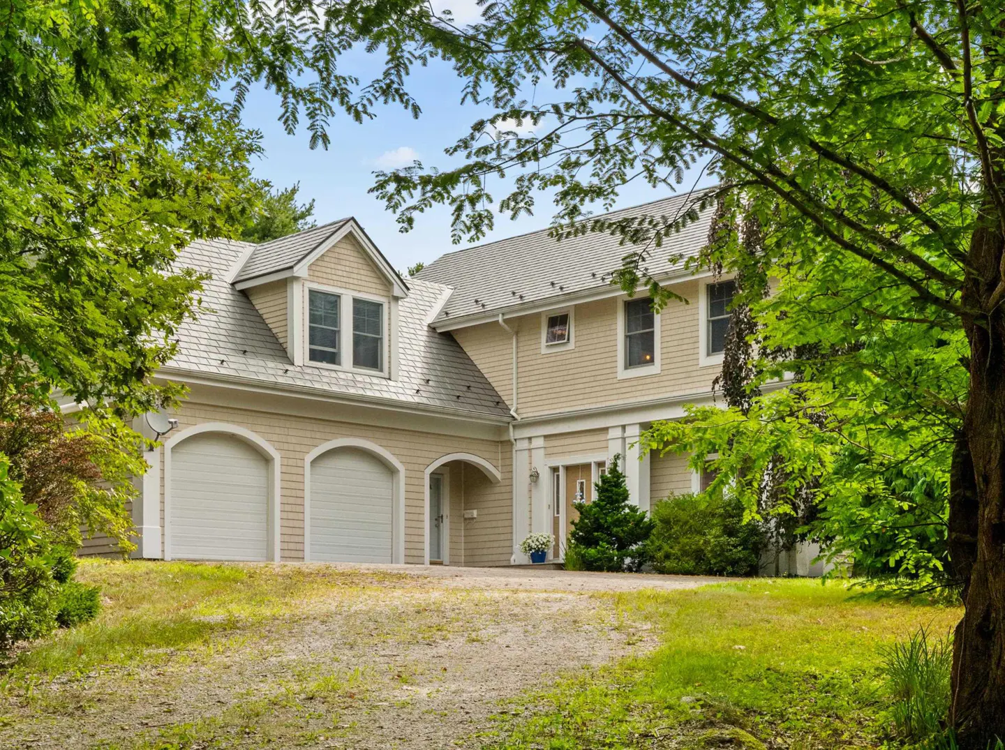Beige two-story house with a gray roof and three-car garage, framed by green trees against a blue sky.