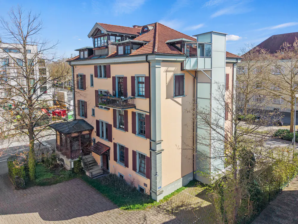 Three-story peach building with red shutters and roof, plus a modern glass elevator shaft.