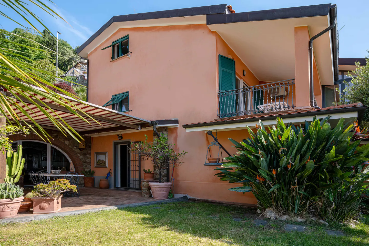 Exterior view of a peach-colored two-story house with green shutters, a balcony, and a patio with a striped awning.