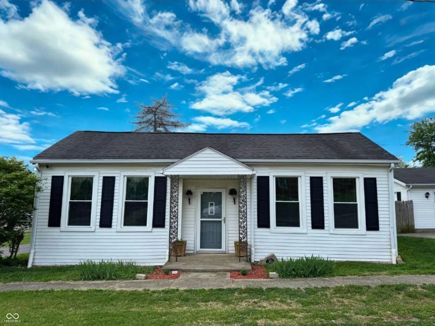 White, one-story house with black shutters and a dark gray roof under a blue sky with white clouds.