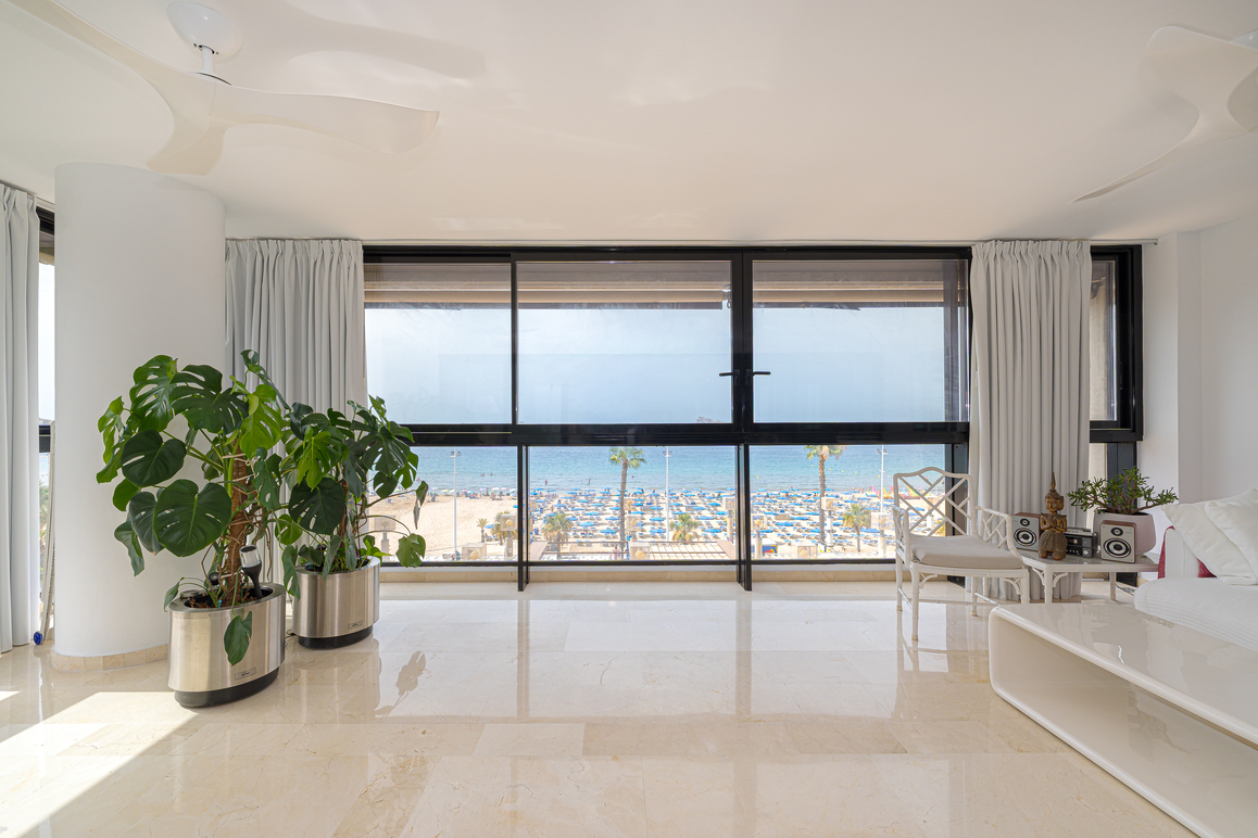Bright living room with large windows overlooking a beach with blue umbrellas, potted plants, and white furniture.