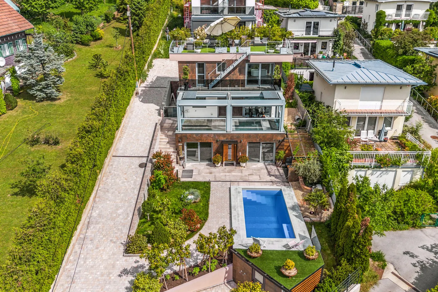 Aerial view of a modern multi-story home with a rooftop terrace, glass-enclosed levels, and a backyard swimming pool.