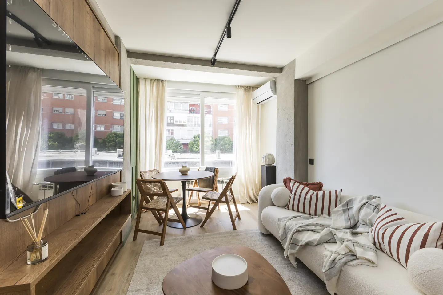 Bright living room with a round table, four chairs, a white sofa with pillows, and a wooden TV stand.