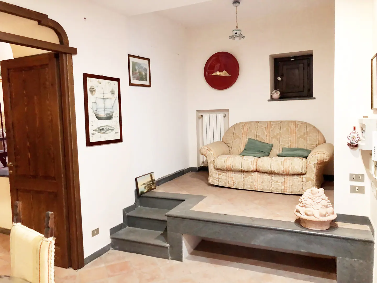 Living room with a beige patterned sofa, green pillows, and gray steps leading to a raised platform. Red plate on the wall.