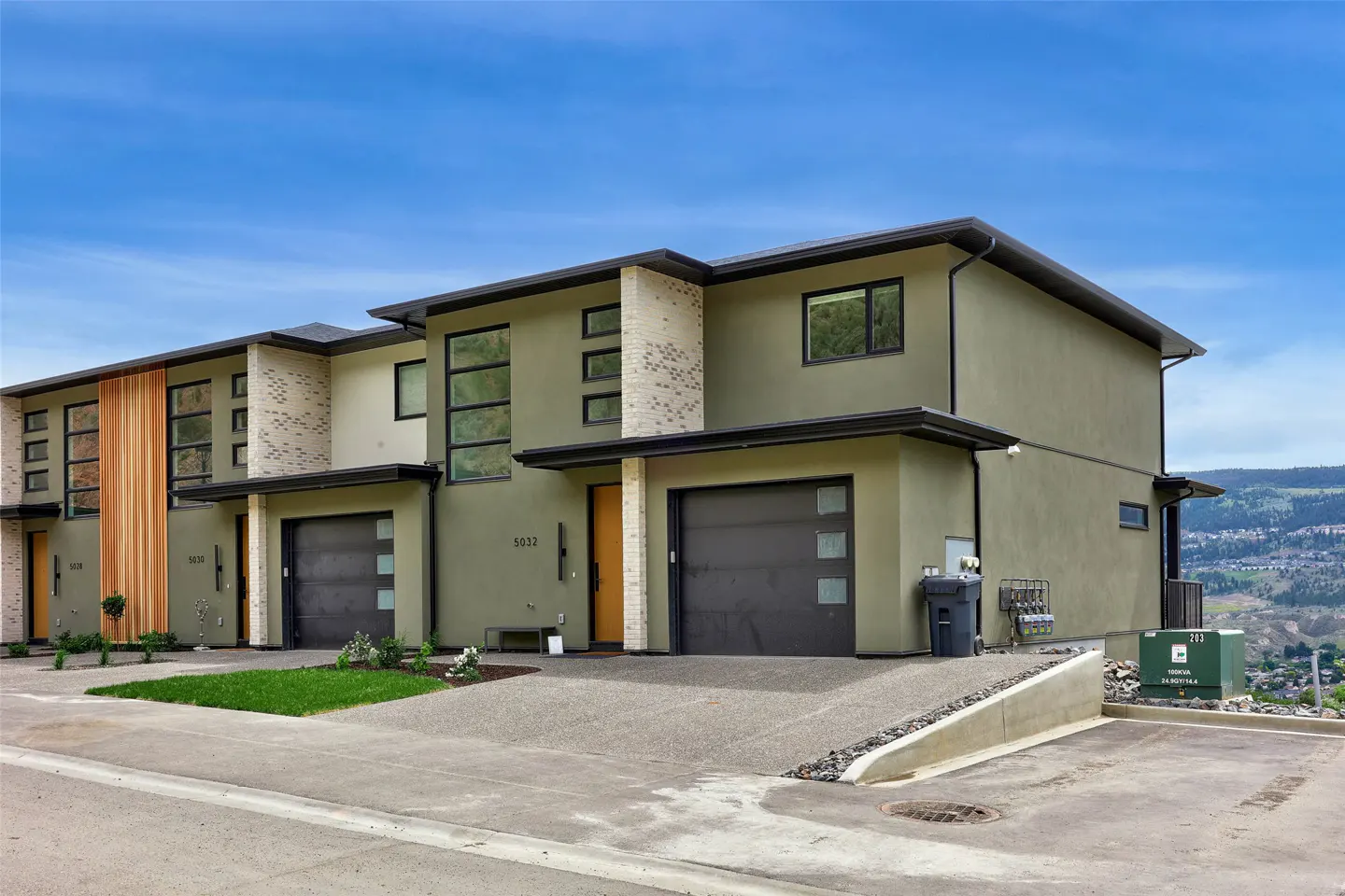 Modern olive-green townhomes with flat roofs, wood accents, and attached garages under a clear blue sky.