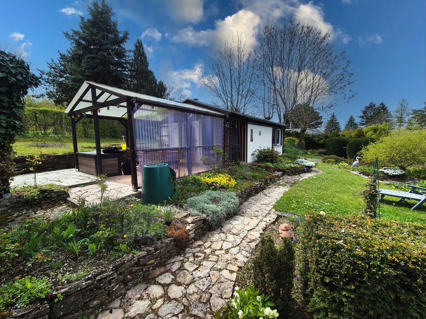 A stone path leads through a lush garden to a white house with a covered patio on a sunny day.