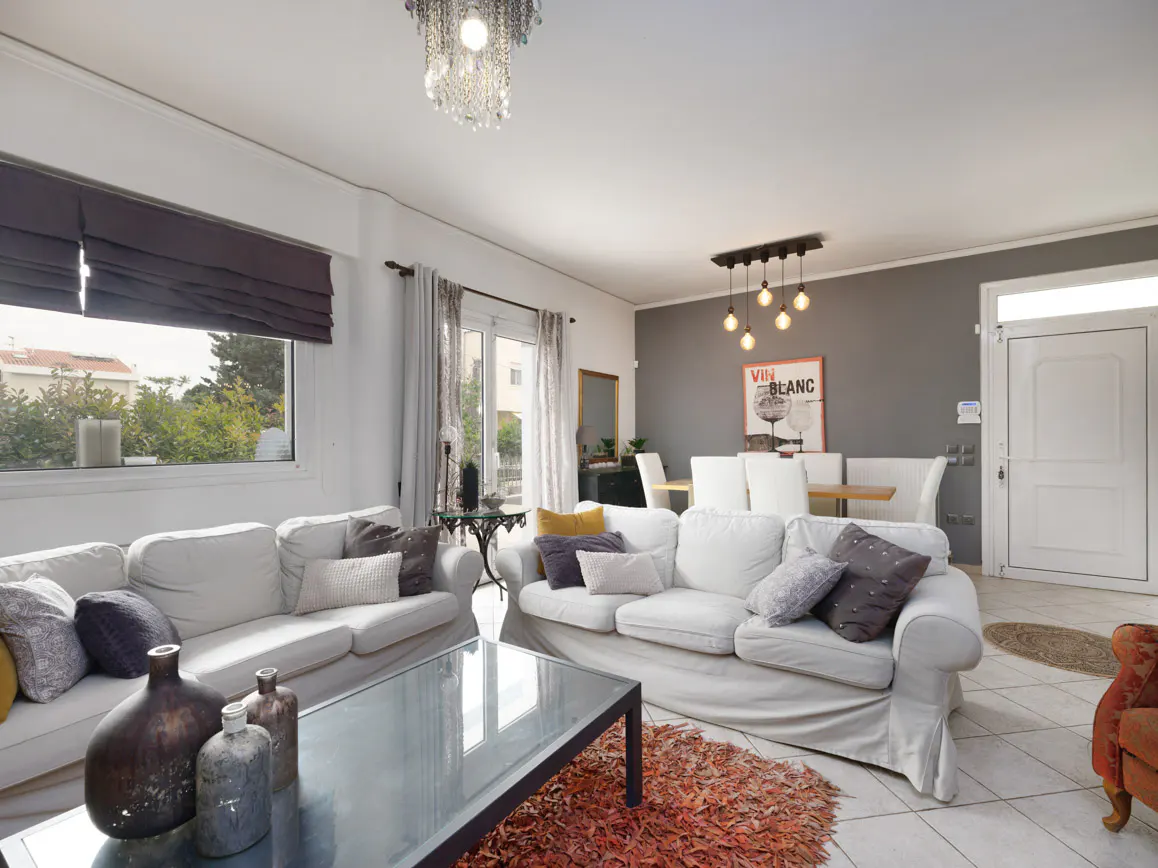 Bright living room with two white sofas, glass coffee table, and dining area with a "Vin Blanc" poster on a gray wall.