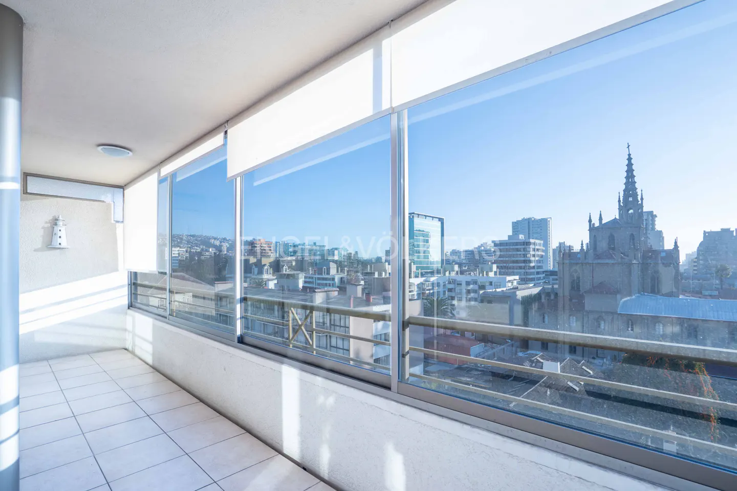 Bright balcony with white tiled floor and glass railings overlooking a city skyline with a prominent church steeple.